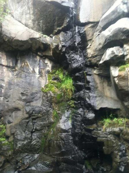 A cascading waterfall flowing down a rocky cliff face, surrounded by dark, textured stones and patches of green vegetation. North Table Mountain mountain bike trail.