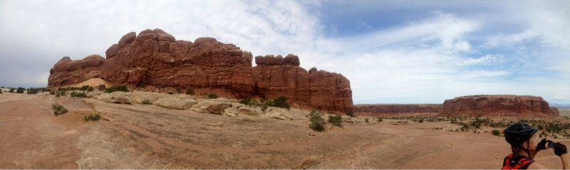 Panoramic view of a desert landscape featuring rugged red rock formations under a partly cloudy sky. A person in cycling gear stands in the foreground, taking a photograph of the scenery. The arid terrain includes patches of sparse vegetation and distant rock cliffs on the horizon. Monitor &amp; Merrimac/Courthouse Pasture mountain bike trail.