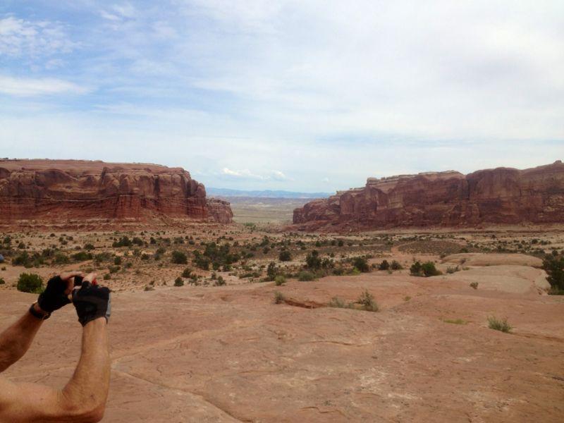 A scenic view of a vast desert landscape featuring large, red rock formations and a cloudy sky. In the foreground, a person's hand is holding a camera, capturing the stunning scenery. Sparse vegetation is visible on the rocky terrain, adding to the natural beauty of the scene. Monitor &amp; Merrimac/Courthouse Pasture mountain bike trail.