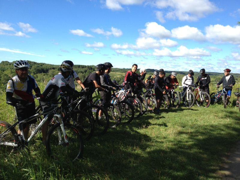 A group of mountain bikers, wearing helmets and cycling gear, stand alongside their bikes on a grassy trail under a clear blue sky. Lush green hills can be seen in the background, suggesting a scenic outdoor environment. The cyclists appear to be preparing for a ride or taking a break, engaged in conversation. Albion Hills mountain bike trail.