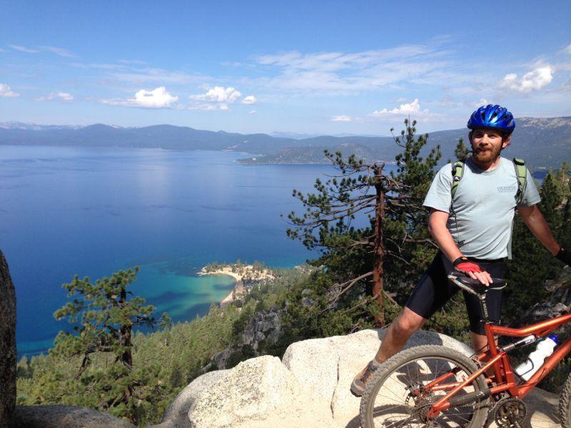 A cyclist in a blue helmet and light blue shirt poses on a rocky trail overlooking a vibrant blue lake surrounded by hills and trees. The sky is clear with a few clouds, and the scenic view showcases a mix of greenery and water. Flume Trail mountain bike trail.