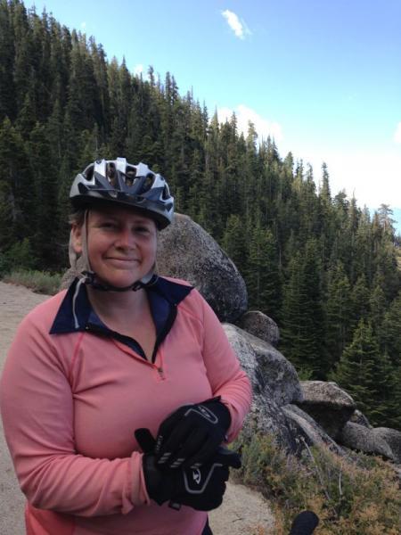 A woman wearing a bike helmet and gloves stands outdoors on a trail. She is smiling and is surrounded by a backdrop of tall evergreen trees and large boulders under a blue sky with scattered clouds. Flume Trail mountain bike trail.