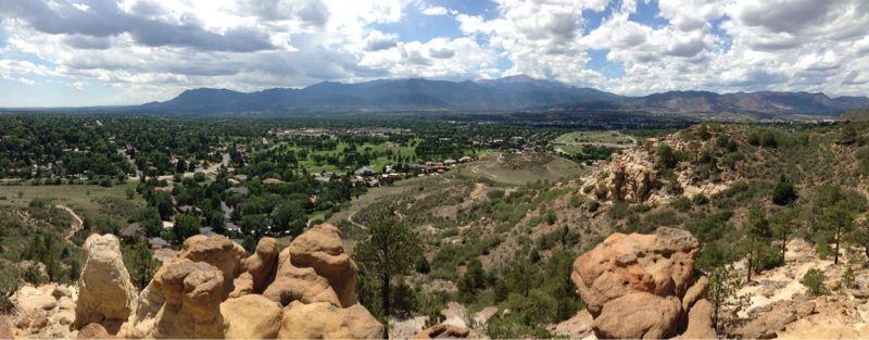 A panoramic view of a lush valley surrounded by mountains, featuring a mix of residential areas and green fields under a partly cloudy sky. Rocky formations are visible in the foreground, and the landscape showcases a blend of natural greenery and developed land. Palmer Park mountain bike trail.