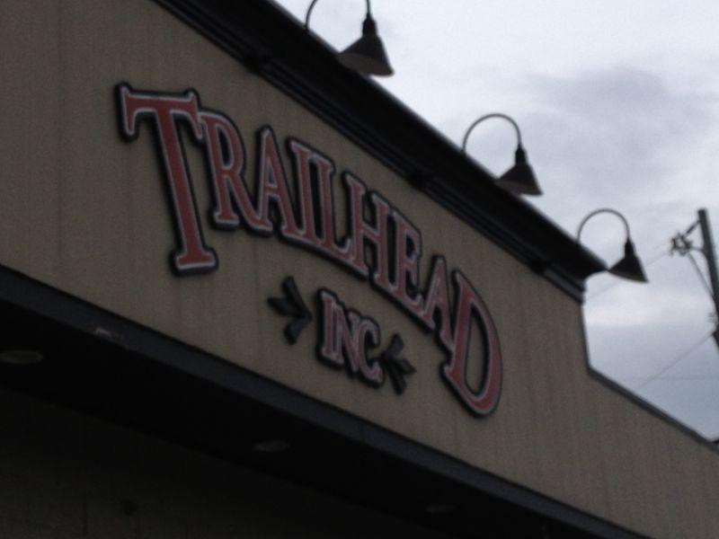Sign for "Trailhead Inc" displayed on the exterior of a building, featuring decorative letters and lighting fixtures above. The sky appears overcast in the background.