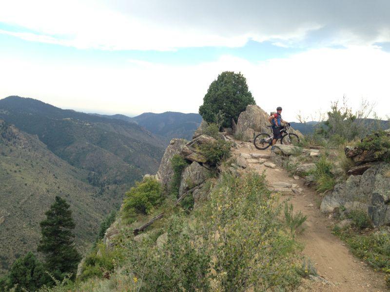 A mountain biker stands next to their bike on a rocky trail overlooking a scenic view of rolling hills and distant mountains, with greenery and shrubs framing the path under a partly cloudy sky. Centennial Cone Park mountain bike trail.