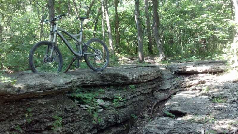 A mountain bike positioned on a rocky outcrop in a lush, green forest. The surrounding trees provide a dappled light effect, highlighting the rugged terrain and the bike's features. A narrow crack in the ground leads away from the bike. Landahl Park Reserve mountain bike trail.