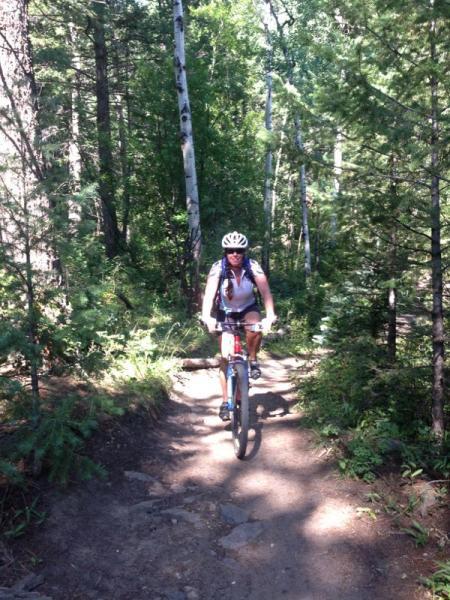 A cyclist wearing a helmet and backpack rides on a dirt trail surrounded by tall trees and greenery in a forested area. The path is narrow and slightly rocky, indicating an outdoor mountain biking adventure. Elk Meadow mountain bike trail.