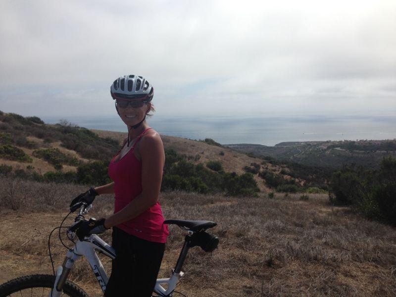 A woman wearing a helmet and sunglasses stands beside a mountain bike on a scenic trail. She is smiling, with rolling hills and a view of the ocean in the background under a cloudy sky. The terrain is grassy and dotted with shrubs. Montana De Oro mountain bike trail.