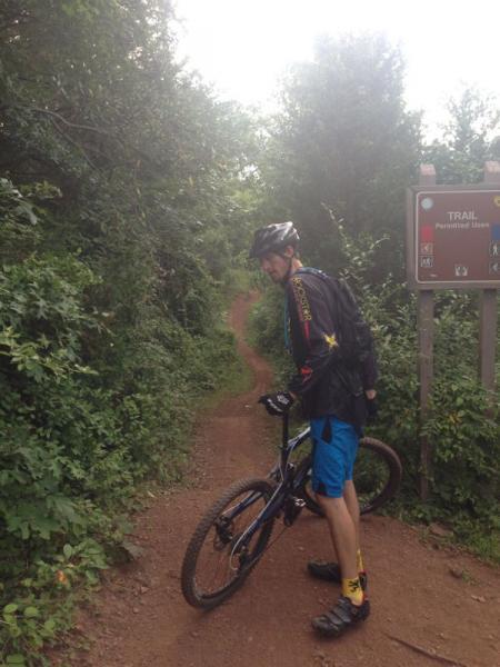 A mountain biker wearing a helmet and cycling gear stands next to a trail sign at the beginning of a dirt path, surrounded by greenery. The biker is holding the handlebars of a blue mountain bike and looking back towards the camera. Six Mile Run mountain bike trail.