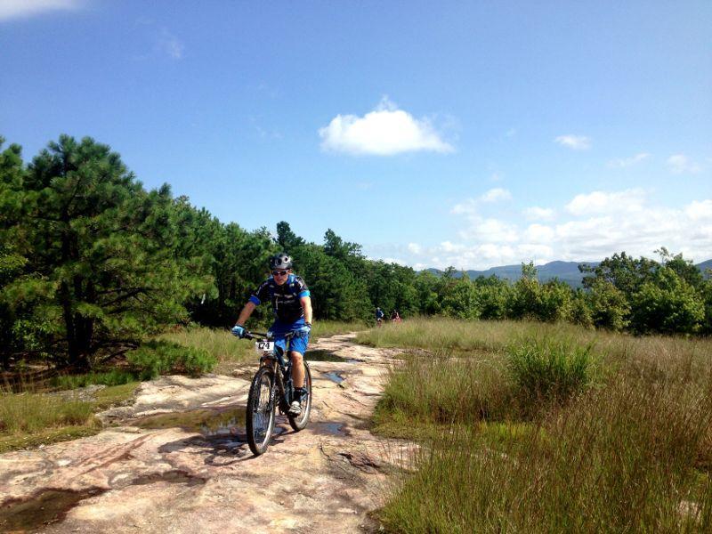 A mountain biker dressed in blue riding along a rocky path surrounded by greenery and trees under a clear blue sky. DuPont State Forest mountain bike trail.