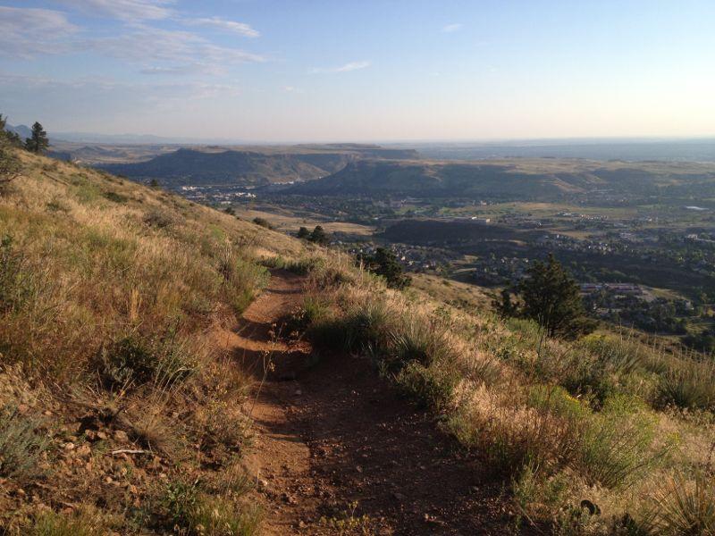 A winding dirt trail leads down a grassy hillside, with a panoramic view of a valley and distant mountains. The landscape features patches of greenery and sparse trees under a blue sky with wispy clouds. The scene captures the tranquility of nature and the expansive beauty of the outdoors. Apex Park mountain bike trail.