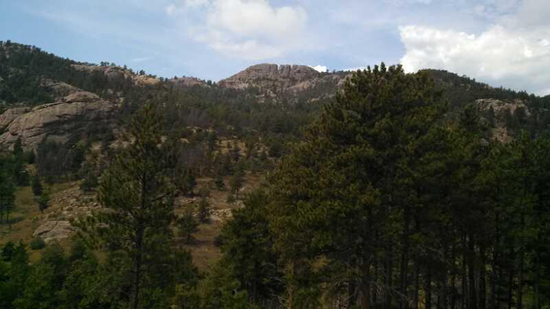 A scenic view of a mountainous landscape featuring rugged hills and dense pine trees under a partly cloudy sky. The foreground includes various evergreen trees, while the background showcases rocky outcrops and peaks. Horsetooth Mountain Park mountain bike trail.