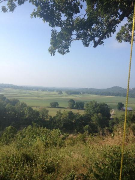 A panoramic view of a lush green valley with rolling hills in the background, captured on a clear day. The foreground features trees and greenery, while the distant hills are silhouetted against the blue sky. A rope is visible on the right side of the image, suggesting an outdoor activity. Bluffview trail mountain bike trail.