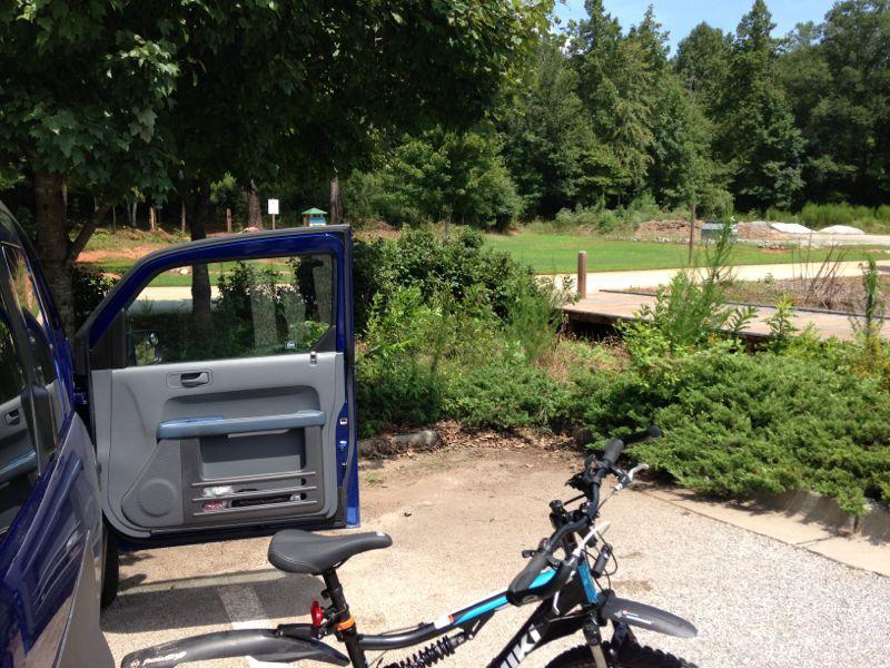 A blue vehicle parked in a shaded area with its front door open. A bicycle is positioned nearby on a gravel surface, with greenery and trees in the background. A pathway and some structures can be seen in the distance, indicating a recreational setting. Yellow River mountain bike trail.