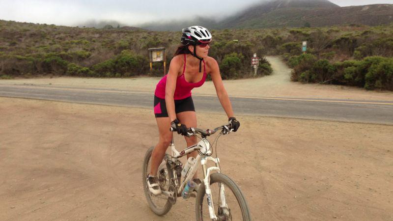 A woman riding a mountain bike on a dirt path, wearing a pink athletic outfit and a helmet. The background features a hilly landscape with low vegetation and a partially cloudy sky. Montana De Oro mountain bike trail.