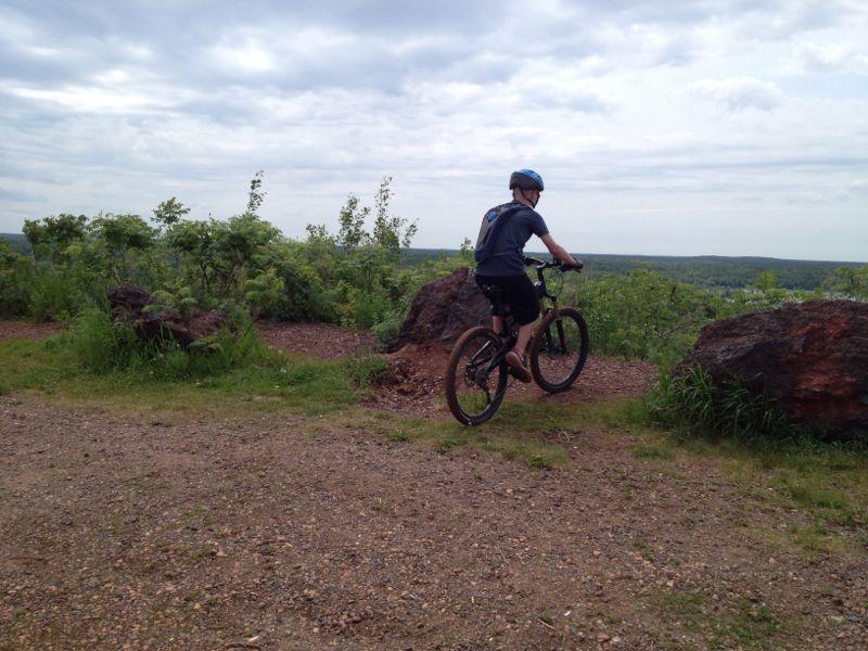 A person on a mountain bike is riding along a scenic dirt path surrounded by greenery and large rocks, overlooking a distant landscape under a partly cloudy sky. Cuyuna Lakes mountain bike trail.
