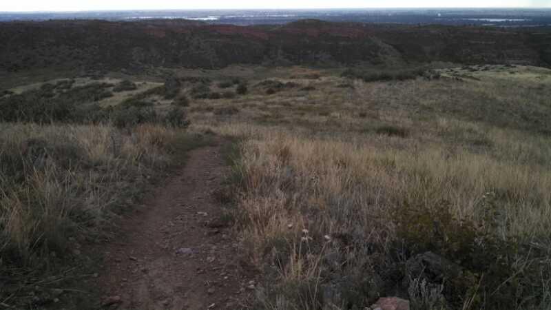A winding dirt path leads through tall, golden grass on a hillside, with a view of rolling hills and a distant body of water under an overcast sky. Indian Summer mountain bike trail.