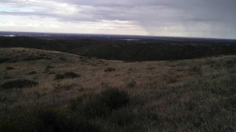 A panoramic view of rolling hills covered in grass, with a cloudy sky above. In the distance, a hint of rain can be seen falling, while the landscape extends to a valley containing trees and possibly water. The scene captures a natural, moody atmosphere typical of a stormy day. Indian Summer mountain bike trail.