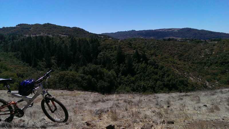 A mountain bike parked on a dry, grassy area overlooking a lush valley filled with trees. In the background, rolling hills and blue sky complete the scenic view. Annadel State Park mountain bike trail.
