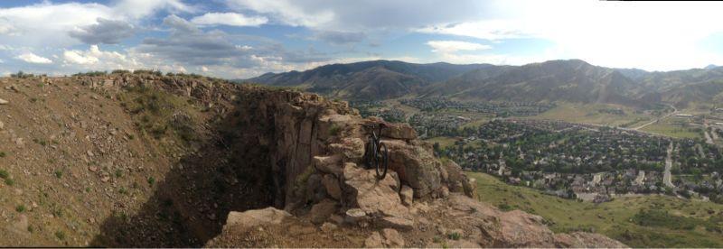 Panoramic view from a rocky cliff overlooking a valley with residential areas and mountains in the background. A bicycle is positioned on the edge of the cliff, suggesting an adventurous outdoor activity. The sky is partly cloudy, creating a dramatic backdrop. North Table Mountain mountain bike trail.