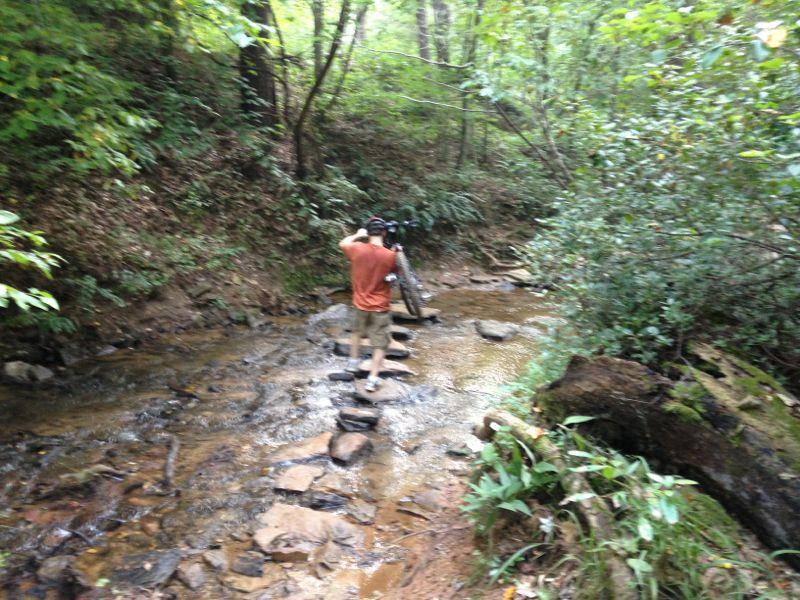 A person walking across stepping stones in a creek, surrounded by lush green vegetation and trees. The individual is carrying a camera on their shoulder, capturing the natural scenery. Chicopee Woods mountain bike trail.