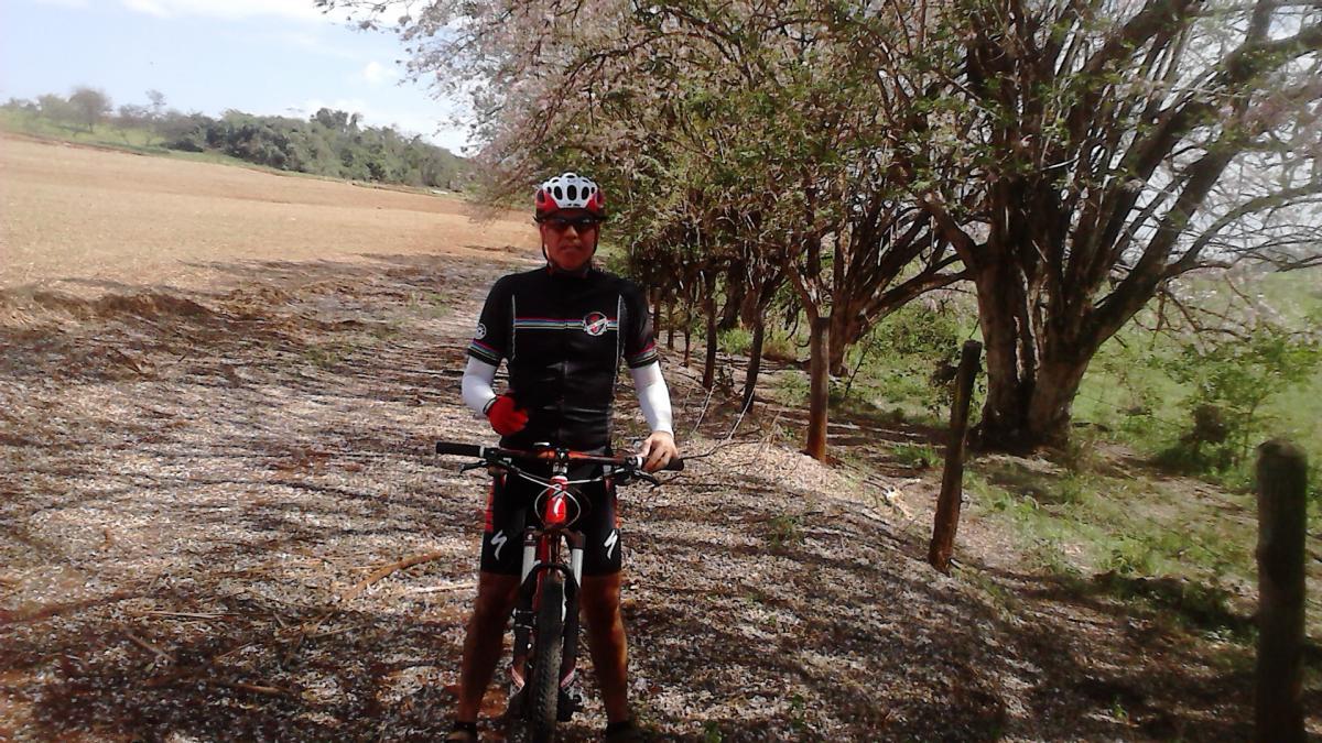 Specialized Epic Comp 29er: An individual wearing a cycling outfit and helmet stands next to a mountain bike on a gravel path surrounded by trees. The background features an open field, and the sky is partly cloudy. The cyclist is making a thumbs-up gesture.