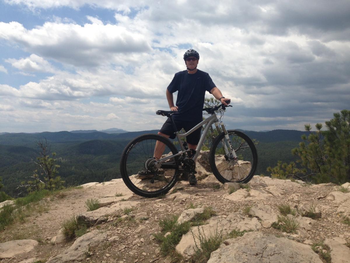 Diamondback Sortie 1: A person standing next to a mountain bike at a scenic viewpoint, surrounded by rolling hills and a cloudy sky. The individual is wearing a helmet and casual cycling attire, with a rugged landscape visible in the background.