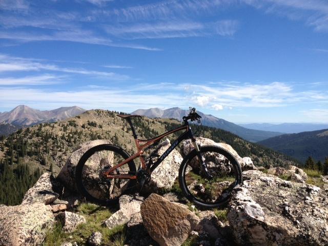 Specialized Epic: A mountain bike resting on rocky terrain overlooking a scenic mountain landscape under a clear blue sky with scattered clouds.