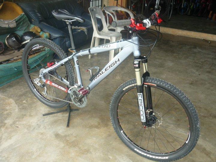 Raleigh Hard tail Mountain Bike: Mountain bike with a silver frame, red and black detailing, and front suspension. The bike is leaning on its side in a garage setting, with visible furniture and additional bikes in the background.