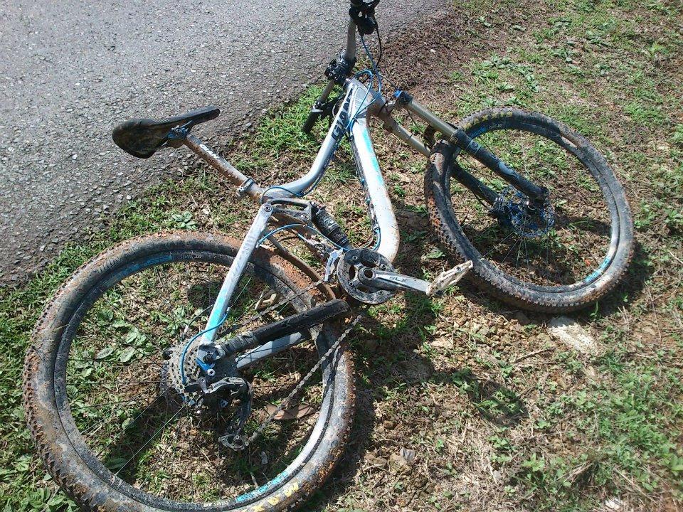 Giant Reign 1: A mountain bike lying on the ground next to a dirt path, partially covered in mud. The bike features a silver and blue frame, with visible dirt on the wheels and components, indicating it has been used off-road. Surrounding the bike is grass and soil, suggesting a natural outdoor setting.