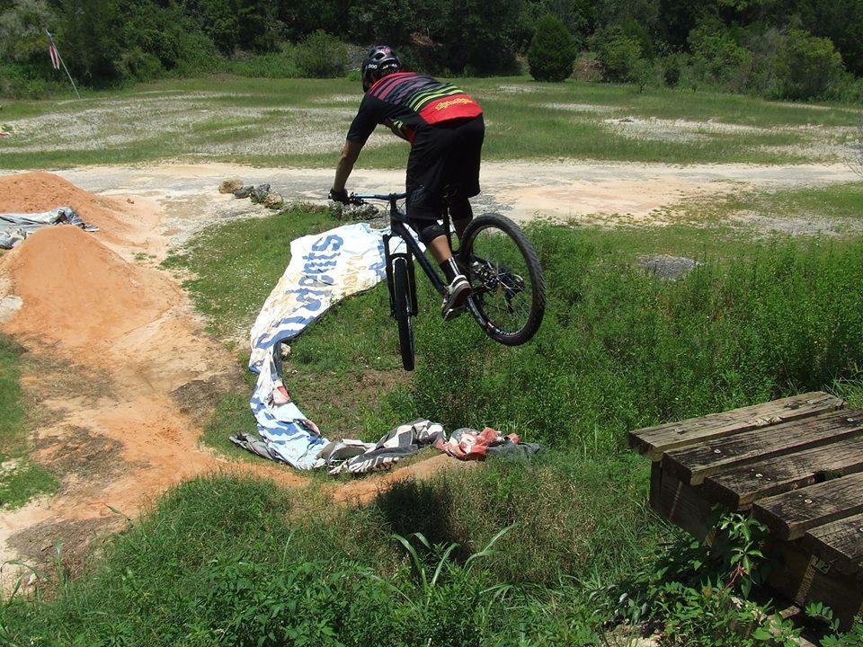 A cyclist wearing a helmet and protective gear jumps off a dirt ramp on a mountain bike, soaring over a tarp with the word "sponsors" visible. The background features a grassy field and trees under clear skies. Santos mountain bike trail.
