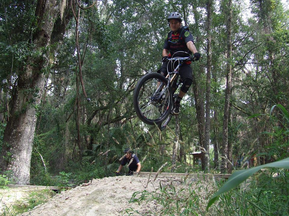 A mountain biker performs a jump on a dirt ramp in a wooded area, surrounded by trees and lush greenery. In the background, a second person watches from near the base of the ramp. The biker is wearing a helmet and protective gear. Santos mountain bike trail.