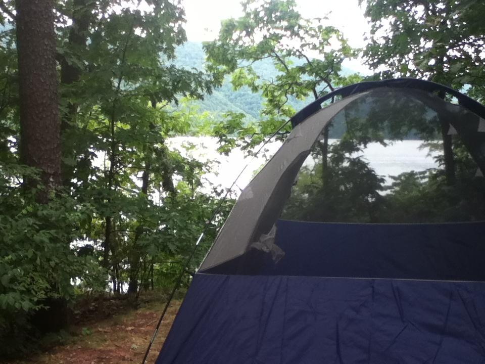 A blue tent set up in a wooded area, with trees surrounding it and a view of a lake in the background. The scene is serene and green, suggesting a peaceful camping experience. Allegrippis Trails mountain bike trail.