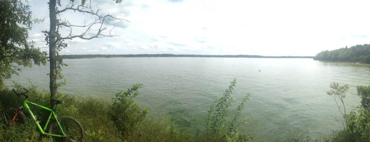 A panoramic view of a calm lake surrounded by lush greenery, with a green bicycle leaning against a tree in the foreground. The sky is partly cloudy, and the shoreline is visible in the distance. Camp Camfield mountain bike trail.