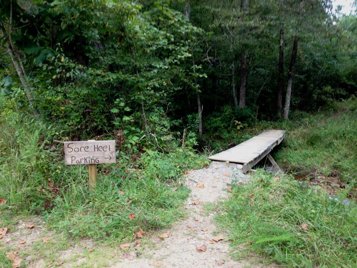 A wooden sign labeled "Sore Heel Parking" points toward a small wooden bridge crossing a grassy path leading into a wooded area. The surroundings are lush with greenery, indicating a natural outdoor setting. Sore Heel Trail mountain bike trail.