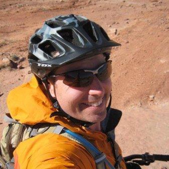 Niner Jet 9 Carbon: A person wearing a black bike helmet and sunglasses smiles at the camera, dressed in an orange jacket, against a backdrop of a sandy terrain.