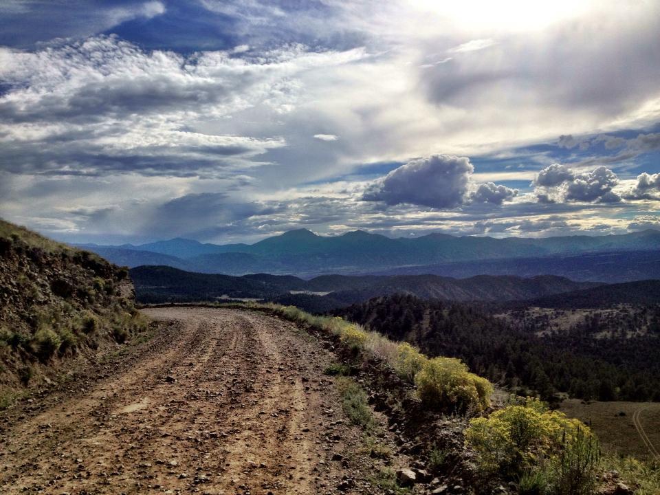 A winding dirt road leads down a hillside, surrounded by lush green vegetation and rocky terrain. In the background, a panoramic view of mountains stretches under a cloudy sky, with layers of blue and gray hues creating a serene landscape. Sunlight breaks through the clouds, casting a soft glow over the scene. CR 175 / Ute Trail mountain bike trail.