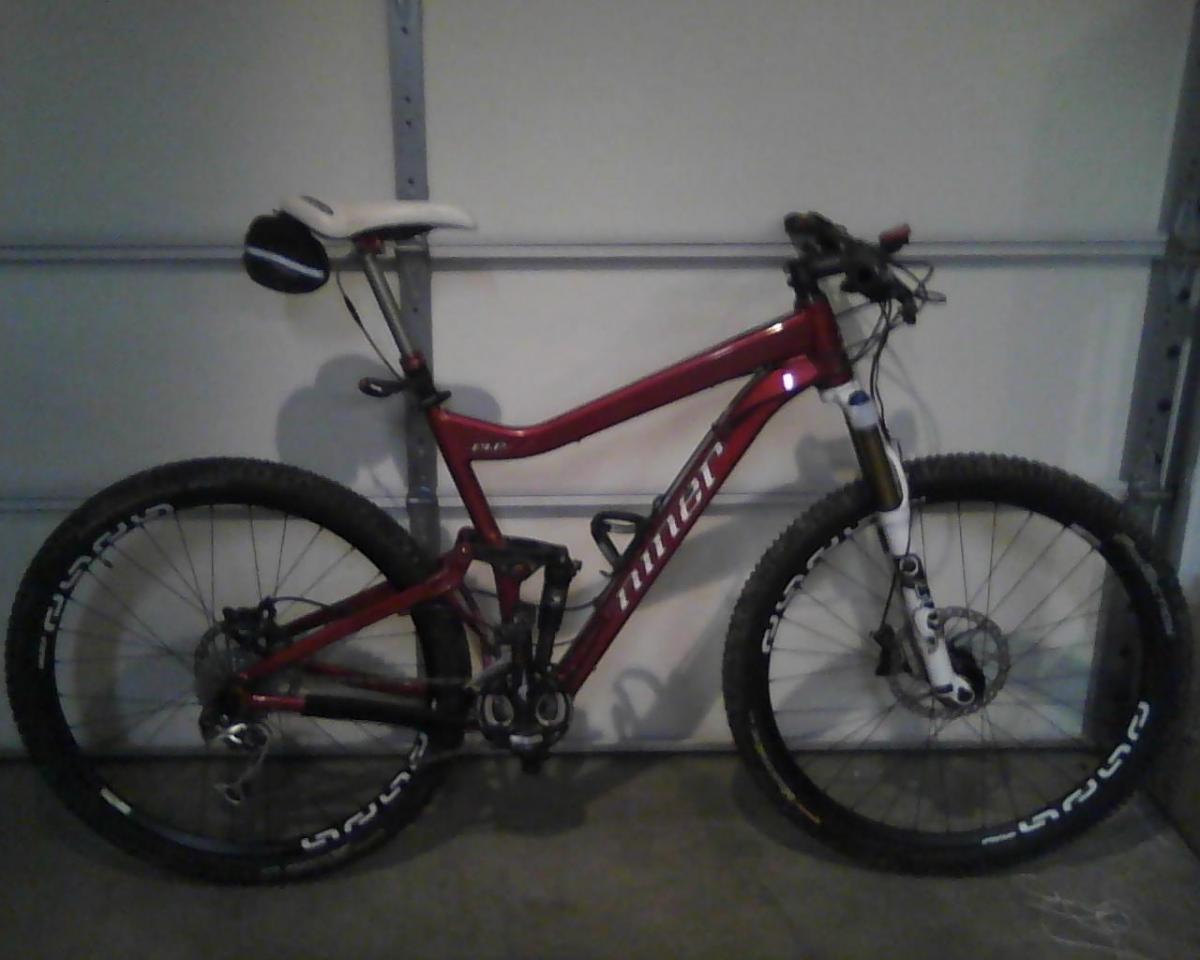 Niner RIP 9: A red mountain bike with a sleek frame, featuring front suspension and wide tires, is positioned against a garage wall. The bike has a white saddle and is equipped with a small bag attached to the seat.