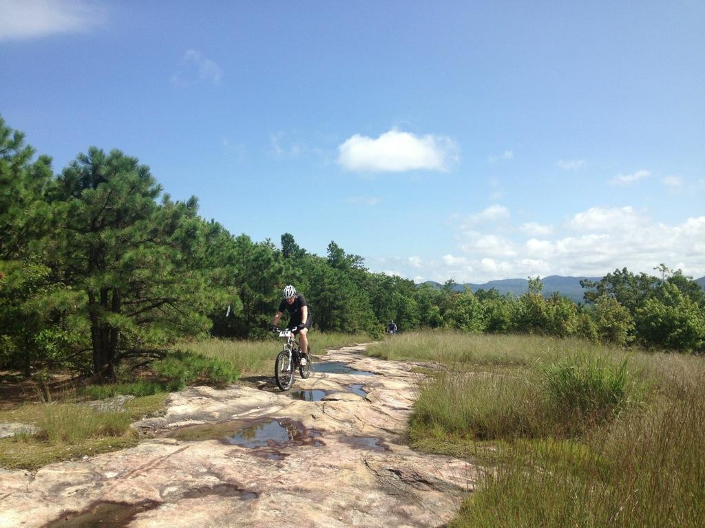 A mountain biker rides along a rocky trail surrounded by greenery and trees under a clear blue sky. The path has visible puddles, and mountains can be seen in the background. Cedar Rock Trail #16 mountain bike trail.