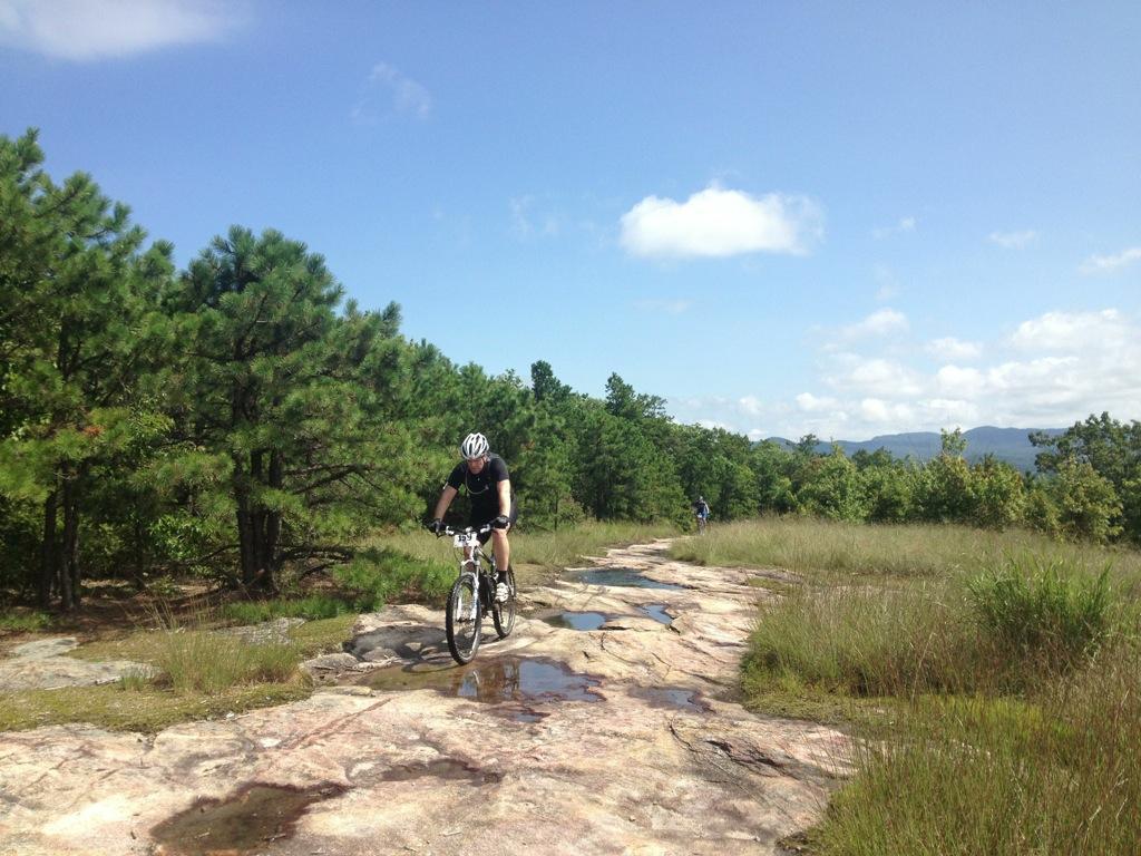 A mountain biker navigating a rocky trail surrounded by trees and grass under a clear blue sky. Puddles of water are visible on the trail, indicating recent rain. Another cyclist can be seen in the background, also riding along the path. Cedar Rock Trail #16 mountain bike trail.