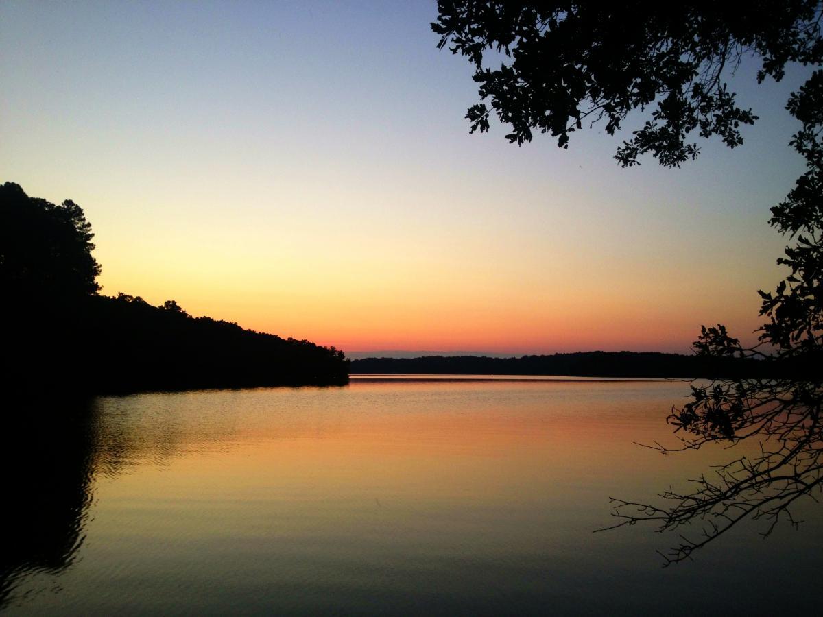 A tranquil lakeside scene at sunset, showcasing a calm water surface reflecting hues of orange, pink, and purple. Silhouetted trees frame the left side of the image against the soft gradient sky. Paynes Creek mountain bike trail.