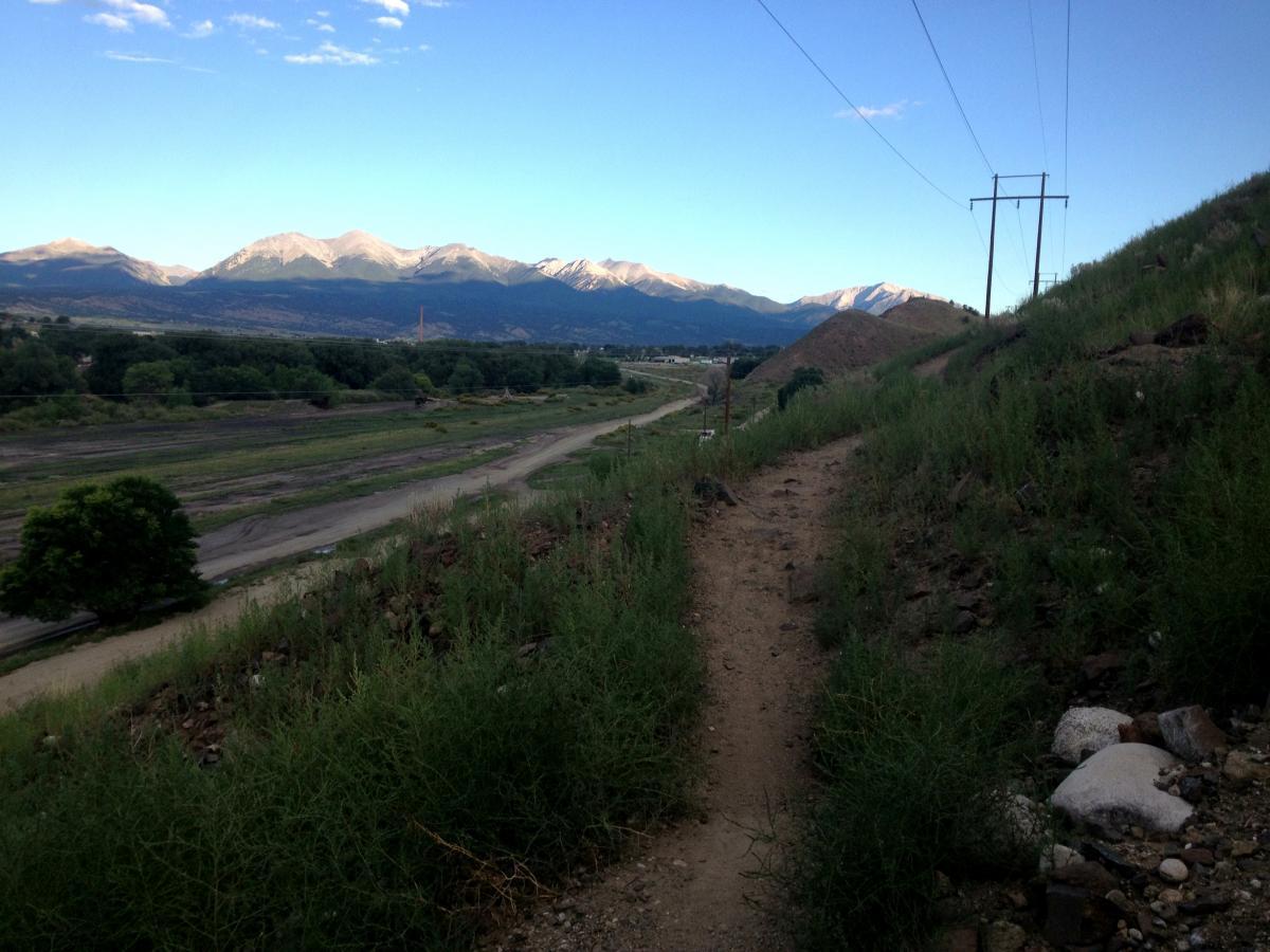A scenic view of a dirt path leading through grassy hills, with mountains in the background under a clear blue sky. The landscape features a mix of greenery and rocky terrain, along with power lines stretching across the horizon. A winding riverbed is visible in the valley below, surrounded by trees and open land. Arkansas Hills mountain bike trail.