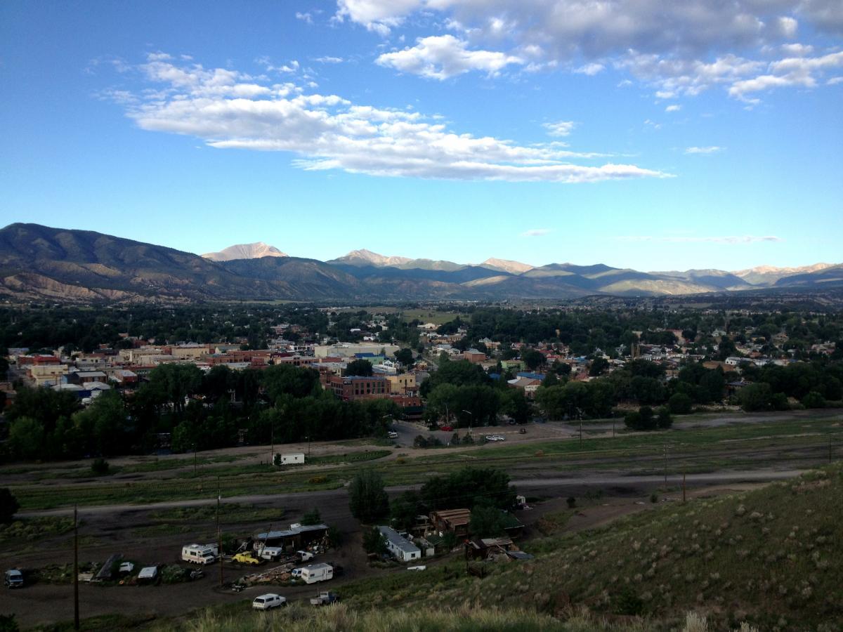 A panoramic view of a scenic valley surrounded by mountains under a blue sky with scattered clouds. In the foreground, a small town is visible with trees and buildings, while the rugged mountains rise majestically in the background. The landscape includes open fields and vehicles parked in the lower area. Arkansas Hills mountain bike trail.
