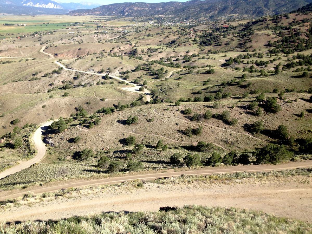 Aerial view of a rugged, hilly landscape with winding dirt roads, sparse vegetation, and scattered trees. In the distance, mountains and a valley can be seen under a clear blue sky. Arkansas Hills mountain bike trail.