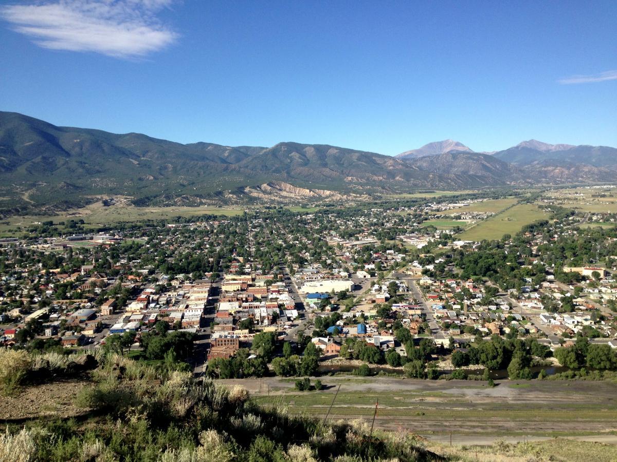 Aerial view of a small town surrounded by green mountains under a clear blue sky. The town features a grid of streets with a mix of residential homes and commercial buildings. Lush green fields and a river are visible in the foreground, while the distant mountains rise majestically in the background. Arkansas Hills mountain bike trail.