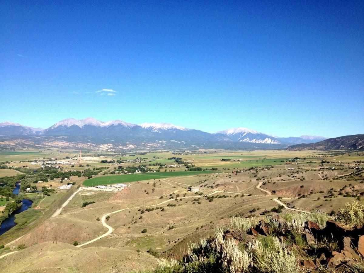 A panoramic view of a mountainous landscape with snow-capped peaks in the background. In the foreground, rolling hills and fields can be seen, along with a winding river and a small town nestled in the valley. The sky is clear and blue, creating a vibrant contrast against the green and brown hues of the land. Arkansas Hills mountain bike trail.