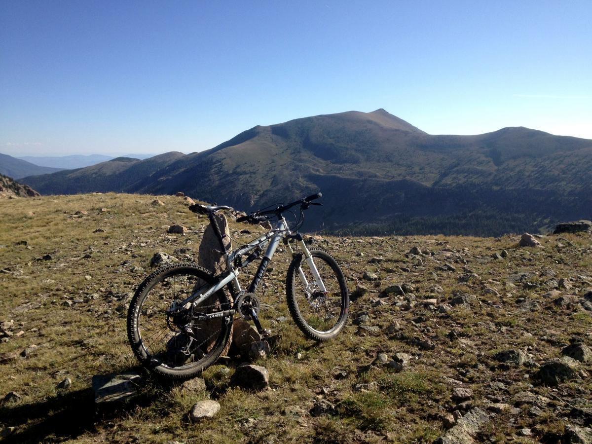 A mountain bike rests against a rock in a grassy field, overlooking a scenic mountain range under a clear blue sky. The landscape features rolling hills and distant peaks, showcasing the beauty of the outdoors. Monarch Crest Trail mountain bike trail.