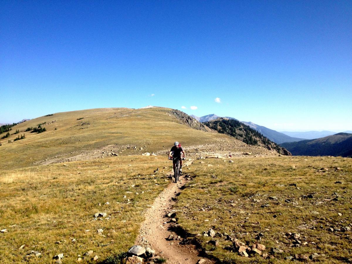 A person riding a mountain bike on a rocky trail in a mountainous landscape, surrounded by rolling hills and a clear blue sky. The vibrant greenery contrasts with the rocky terrain and distant peaks. Monarch Crest Trail mountain bike trail.