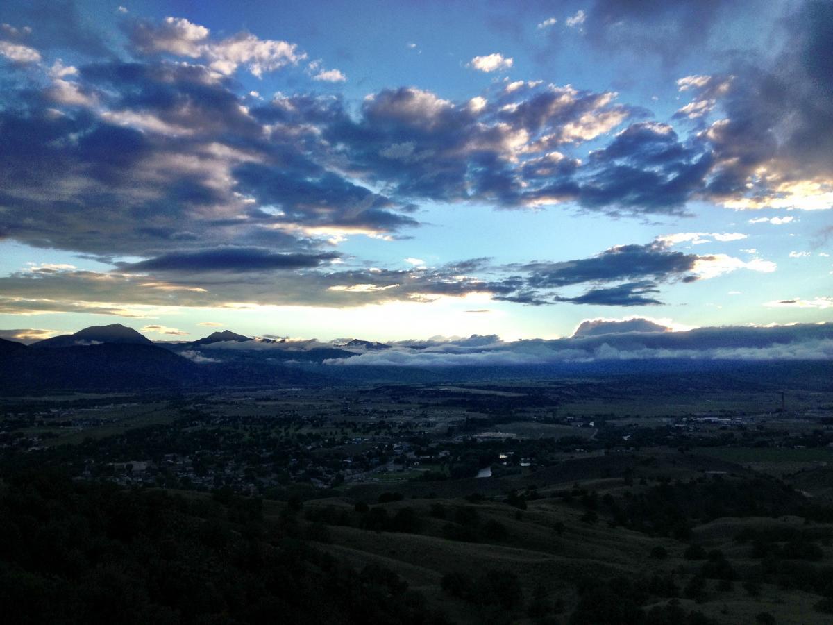 A scenic view of a valley surrounded by mountains under a partly cloudy sky at sunset. The clouds exhibit shades of blue and gray, while the landscape features rolling hills and patches of greenery. The horizon showcases the silhouettes of distant mountains, partially covered by mist. Arkansas Hills mountain bike trail.