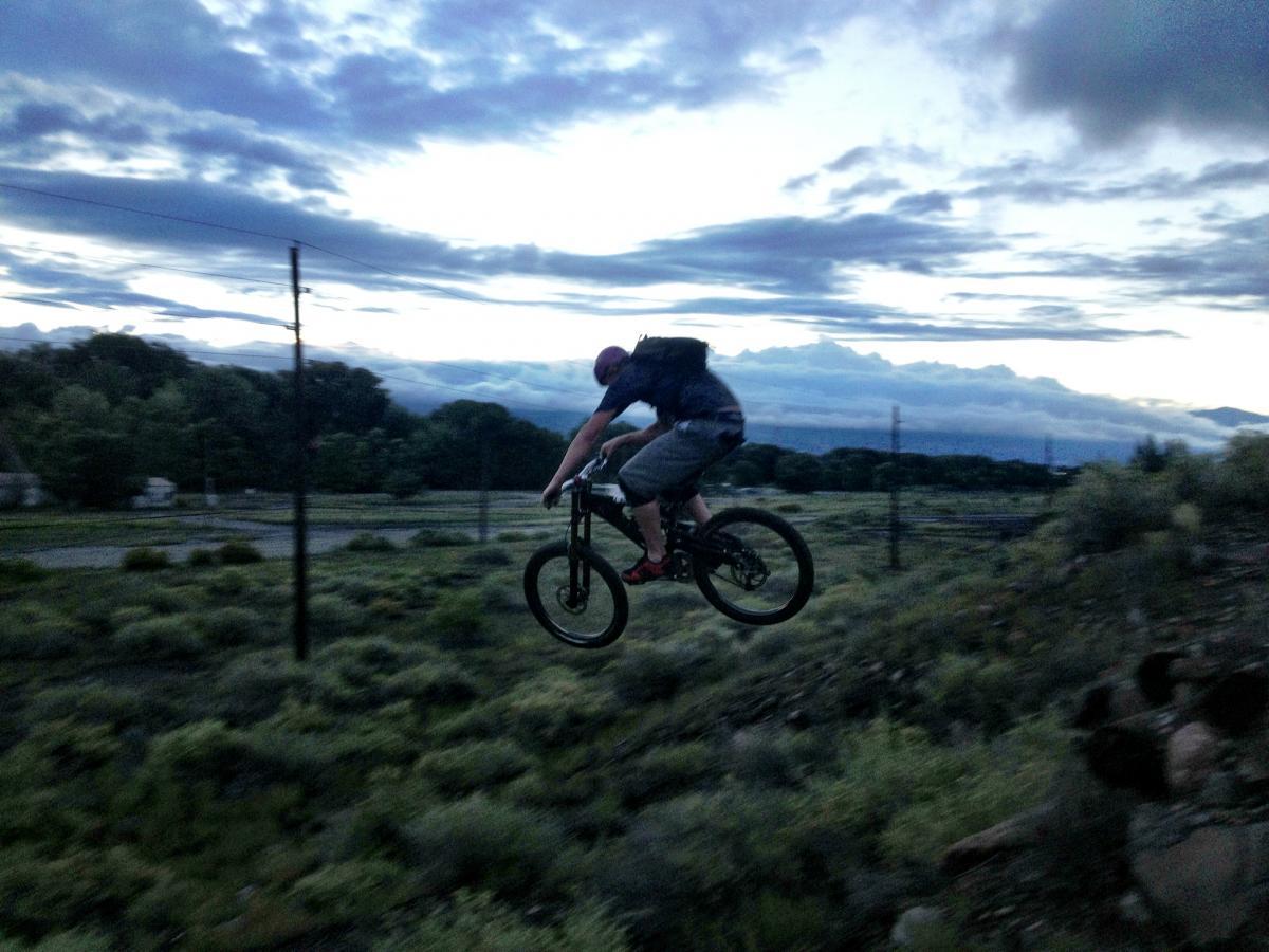 A mountain biker in mid-air executing a jump over a grassy hill at dusk, with lush greenery and a cloudy sky in the background. Arkansas Hills mountain bike trail.